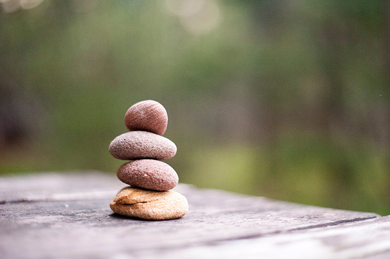 f29dbdcbd38cbc003f4470618cb58d39.jpg Four pebbles stacked onto of each other on a flat surface. The background is blurred and green.